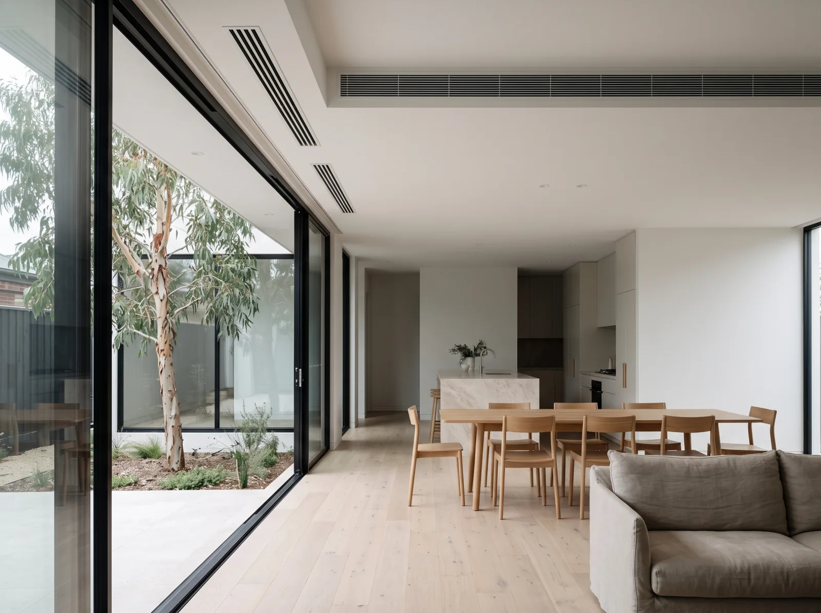 Open-plan living, dining and kitchen with pale oak floor, black-framed sliding glass doors open to a courtyard with a gum tree, an oak dining table and a stone island. A long linear supply grille sits in a recessed ceiling bulkhead, with two smaller slot grilles above the glazing.