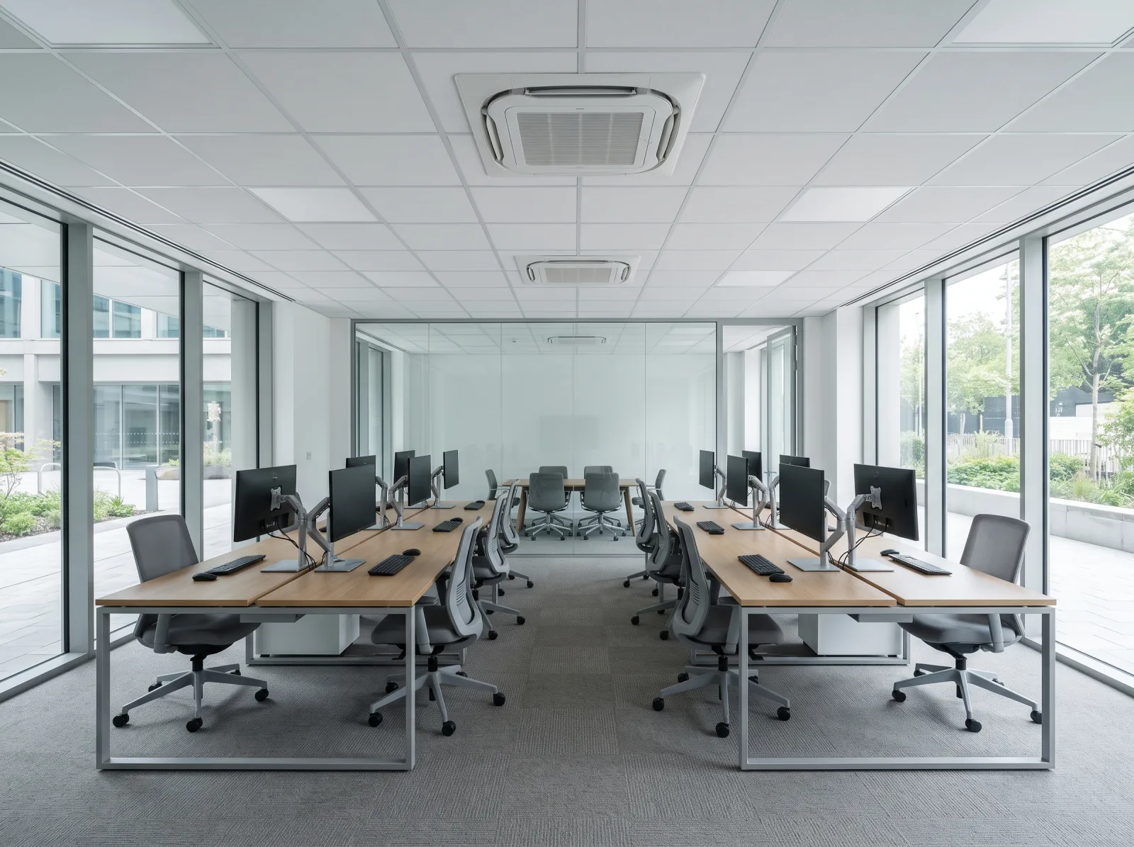 Open-plan office with two rows of oak desks and grey task chairs on grey carpet tiles, full-height glazing on both sides and a glass-walled meeting room at the far end. Two four-way ceiling cassette units sit flush in the white suspended ceiling grid, with a third visible inside the meeting room.